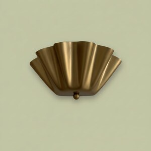 Könnte beinhalten: Eine goldfarbene Wandleuchte mit einem wellenförmigen, fächerartigen Design. Die Leuchte hat ein kleines, rundes Detail an der Unterseite. Die Leuchte ist aus Metall und an einer hellgrünen Wand befestigt.