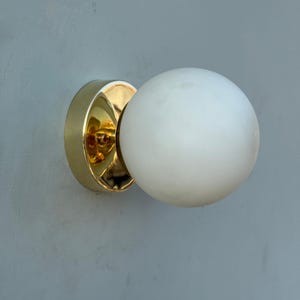 May include: A gold-colored wall sconce with a white, spherical light fixture. The sconce has a circular base and a reflective, polished finish. The light fixture is a smooth, opaque sphere.