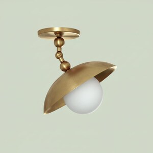 Könnte beinhalten: Eine Deckenleuchte in Messingoptik mit einer kugelförmigen, weißen Glühbirne. Die Leuchte hat einen geschwungenen Schirm und einen verstellbaren Arm und ist auf einer kreisförmigen Basis montiert.