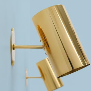 Könnte beinhalten: Zwei goldfarbene Wandleuchten mit zylindrischem Schirm und runder Montageplatte. Die Leuchten sind an einer hellblauen Wand montiert. Die Leuchten haben ein modernes, minimalistisches Design.
