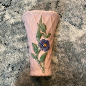 May include: A pink ceramic vase with a textured diamond pattern. The vase is decorated with a blue flower and green leaves. The vase is approximately 6 inches tall.