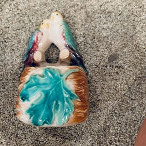 May include: A ceramic figurine featuring two birds perched on a branch. The birds have white, yellow, and teal plumage with red accents. The base is brown with a teal leaf design.