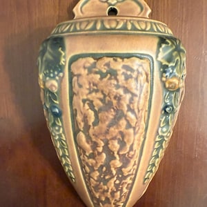 May include: A vintage wall pocket vase in a muted color palette. The vase features a textured, central panel and is adorned with relief details of leaves and berries. The top has a decorative, arched design with a hole for hanging.