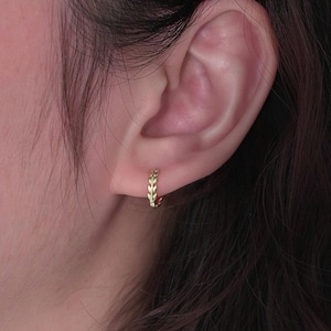 Puede incluir: Primer plano de un pendiente de aro dorado con diseño de hojas. El pendiente se lleva en una oreja, con cabello castaño oscuro en el fondo. El pendiente es pequeño y delicado, con un patrón repetido de hojas.