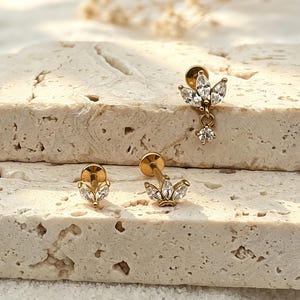 Può includere: Tre orecchini a lobo color oro con pietre trasparenti taglio marquise disposte a forma di foglia. Un orecchino include una pietra rotonda trasparente pendente. Ogni pezzo presenta una chiusura circolare piatta e un sottile perno in metallo.