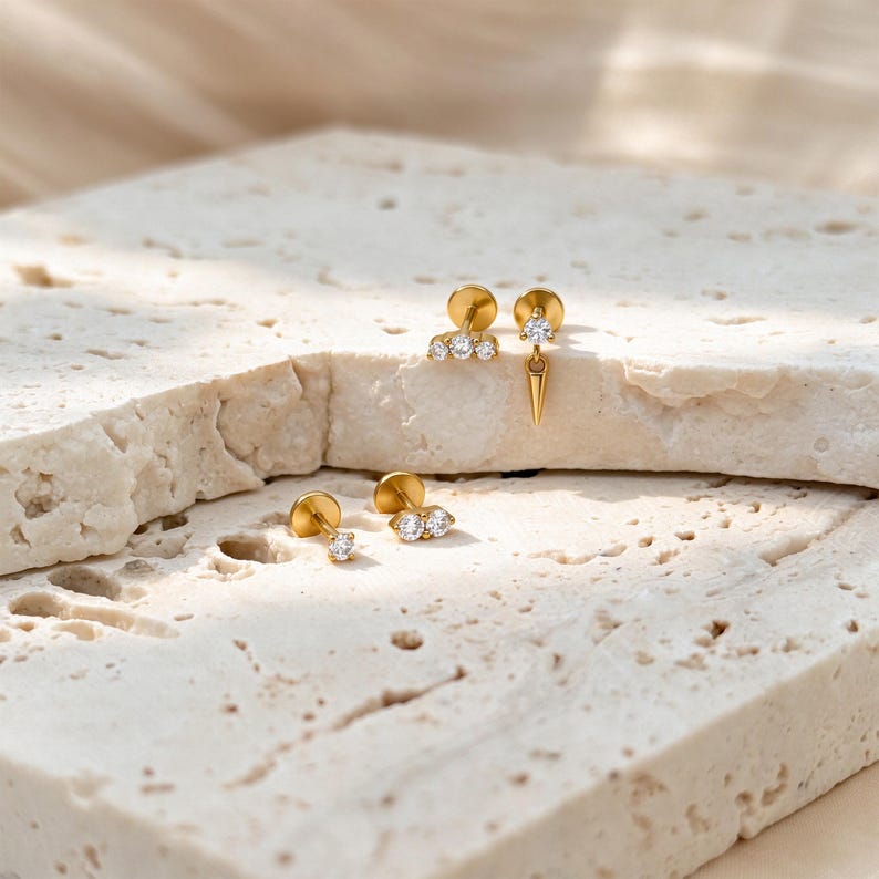 May include: Four gold-tone stud earrings featuring clear, round-cut stones. One earring has a three-stone cluster, one has a single stone with a dangling spike, one has a single stone, and one has a two-stone cluster. All have flat backings.