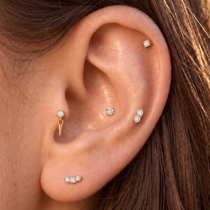 May include: A collection of gold-toned earrings featuring clear, round-cut stones. The set includes a single stud, a double-stone stud, a triple-stone curved stud, and a stud with a dangling gold spike charm, all worn in various ear piercings.