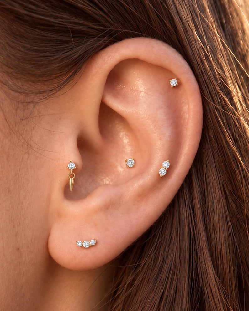 May include: A collection of gold-toned earrings featuring clear, round-cut stones. The set includes a single stud, a double-stone stud, a triple-stone curved stud, and a stud with a dangling gold spike charm, all worn in various ear piercings.