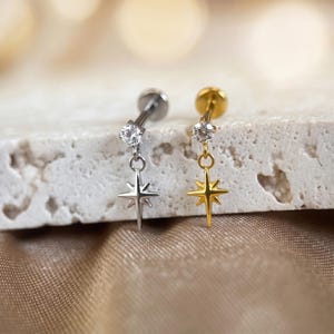 May include: Two star-shaped earrings, one silver-toned and one gold-toned, each with a dangling star and a small, clear gemstone. The earrings are displayed against a neutral, textured background, showcasing their delicate design and contrasting colors.