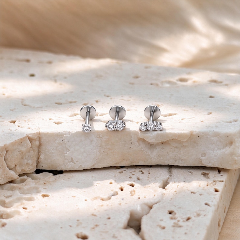 May include: Three silver-toned flat back stud earrings with clear round stones. The first earring features a single stone, the second has two stones side-by-side, and the third displays three stones in a row, all with a metallic post and disc backing.