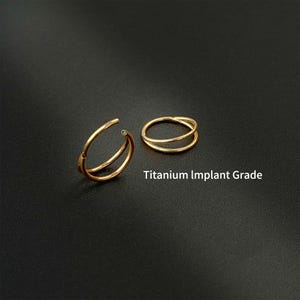 Op de afbeelding: Twee goudkleurige neusringen met dubbele ringen op een zwarte achtergrond. Eén ring is open en toont de sluiting, de andere is gesloten. De tekst "Titanium Implant Grade" is in het wit gedrukt.