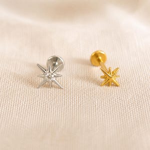 Könnte beinhalten: Zwei sternförmige Ohrstecker. Einer ist silberfarben mit einem klaren Edelstein in der Mitte, der andere ist goldfarben. Beide Ohrringe haben einen Stecker und einen Verschluss. Die Ohrringe liegen auf einem cremefarbenen Stoff.