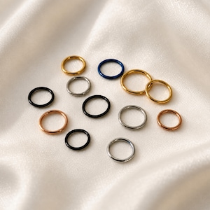 May include: A collection of eleven circular hoop rings in various metallic finishes including gold, silver, rose gold, black, and blue. Each ring features a simple, thin, continuous band design with a seamless closure mechanism visible on several pieces.