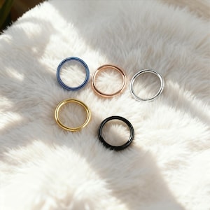 Puede incluir: Cinco anillos metálicos en varios colores: azul, oro rosa, plata, oro y negro. Los anillos son circulares y parecen ser de metal. Están dispuestos sobre una superficie suave, blanca y esponjosa.