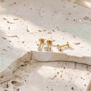 May include: Three gold-toned flat back stud earrings with clear, round-cut stones. One earring features a single stone, while the other two earrings each feature a pair of stones set side-by-side on a small metal bar, all with thin posts.
