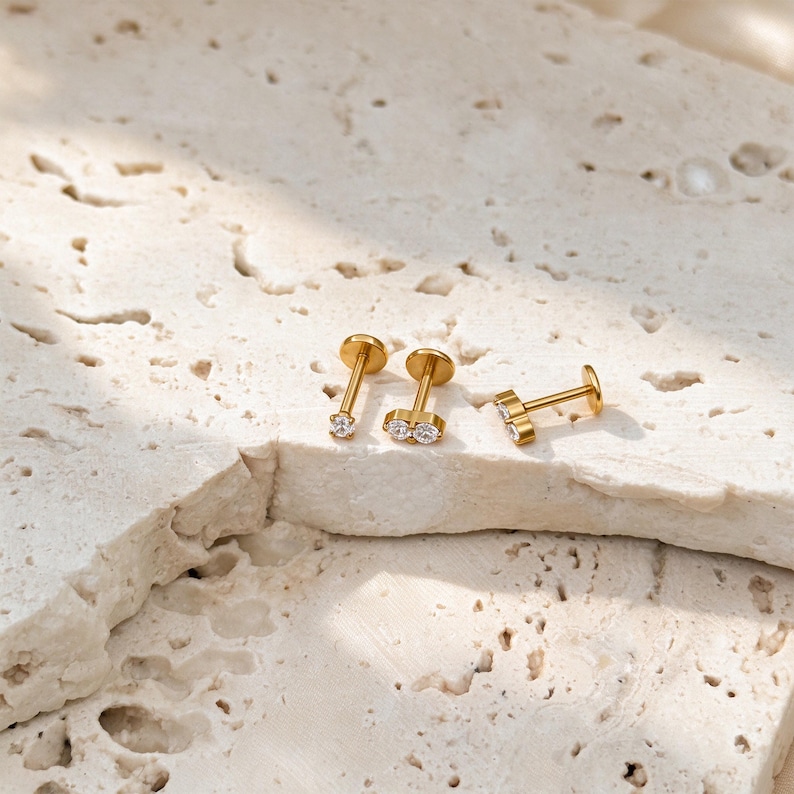 May include: Three gold-toned flat back stud earrings with clear, round-cut stones. One earring features a single stone, while the other two earrings each feature a pair of stones set side-by-side on a small metal bar, all with thin posts.