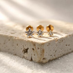 Puede incluir: Tres pendientes de botón en tono dorado con un diseño en forma de estrella y una piedra preciosa transparente y facetada en el centro. Los pendientes varían en tamaño, cada uno con un cierre circular plano y un mecanismo de poste seguro.