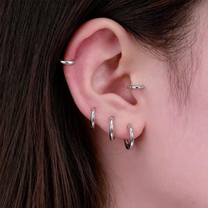 May include: Close-up of an ear with multiple silver hoop earrings and a silver ear cuff. The earrings vary in size, with three small hoops in the earlobe and a larger hoop in the upper ear. The ear cuff is a simple, thin band.