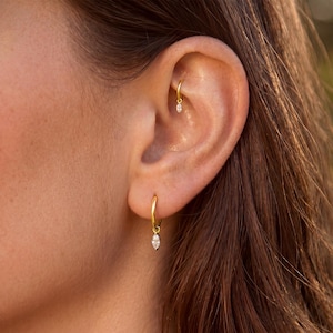Könnte beinhalten: Nahaufnahme eines Ohrs mit zwei goldenen Creolen. Der größere Ohrring hat einen hängenden, marquiseförmigen Edelstein. Ein kleinerer Creolen-Ohrring mit einem ähnlichen Edelstein befindet sich im oberen Ohr. Die Ohrringe haben eine polierte, reflektierende Oberfläche.