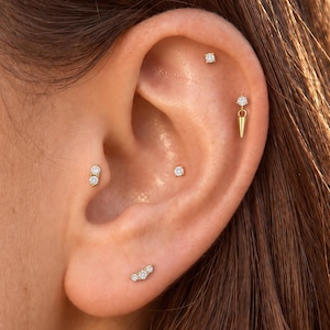 May include: A collection of gold earrings featuring clear round stones. The set includes a three-stone cluster stud, a two-stone vertical stud, two single-stone studs, and a single-stone stud with a dangling gold spike charm.