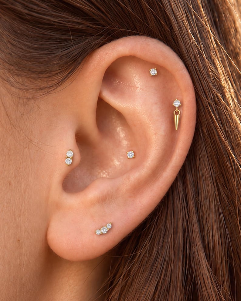 May include: A collection of gold earrings featuring clear round stones. The set includes a three-stone cluster stud, a two-stone vertical stud, two single-stone studs, and a single-stone stud with a dangling gold spike charm.