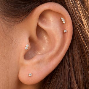 May include: A set of four gold-tone earrings featuring clear, round-cut stones. One earring is a single stone in the lobe, one is a two-stone cluster in the tragus, and two are positioned in the upper ear cartilage, one with three stones and one single.