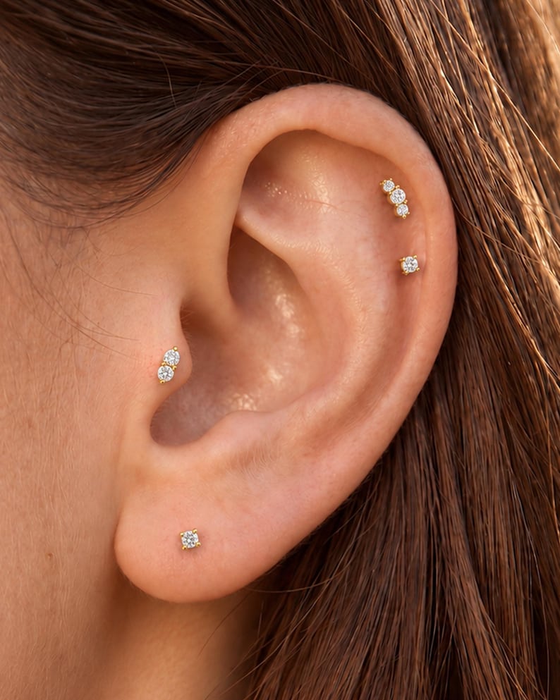 May include: A set of four gold-tone earrings featuring clear, round-cut stones. One earring is a single stone in the lobe, one is a two-stone cluster in the tragus, and two are positioned in the upper ear cartilage, one with three stones and one single.