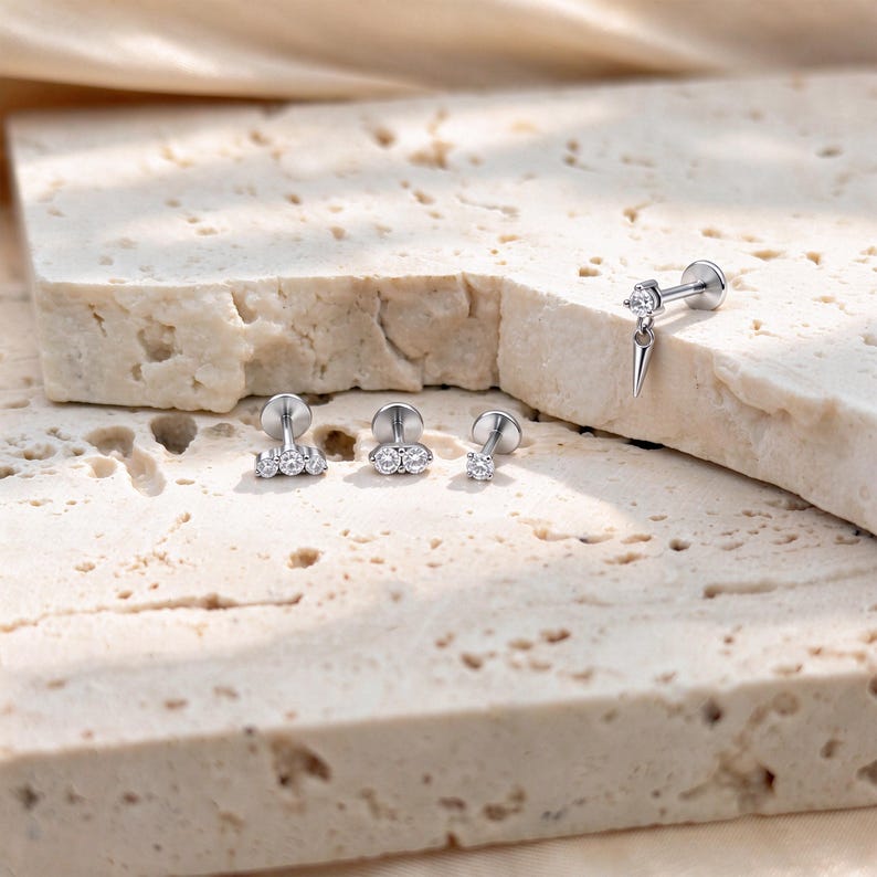 May include: Four silver-toned metal stud earrings with clear stones. One earring features a dangling spike charm, one has three stones in a row, one has two stones side-by-side, and one is a single stone stud. Each piece has a flat circular back.