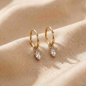 Puede incluir: Un par de pendientes de aro dorados con una gema transparente en forma de marquesa colgante. Los pendientes se exhiben sobre una superficie beige satinada, creando una presentación lujosa y elegante.
