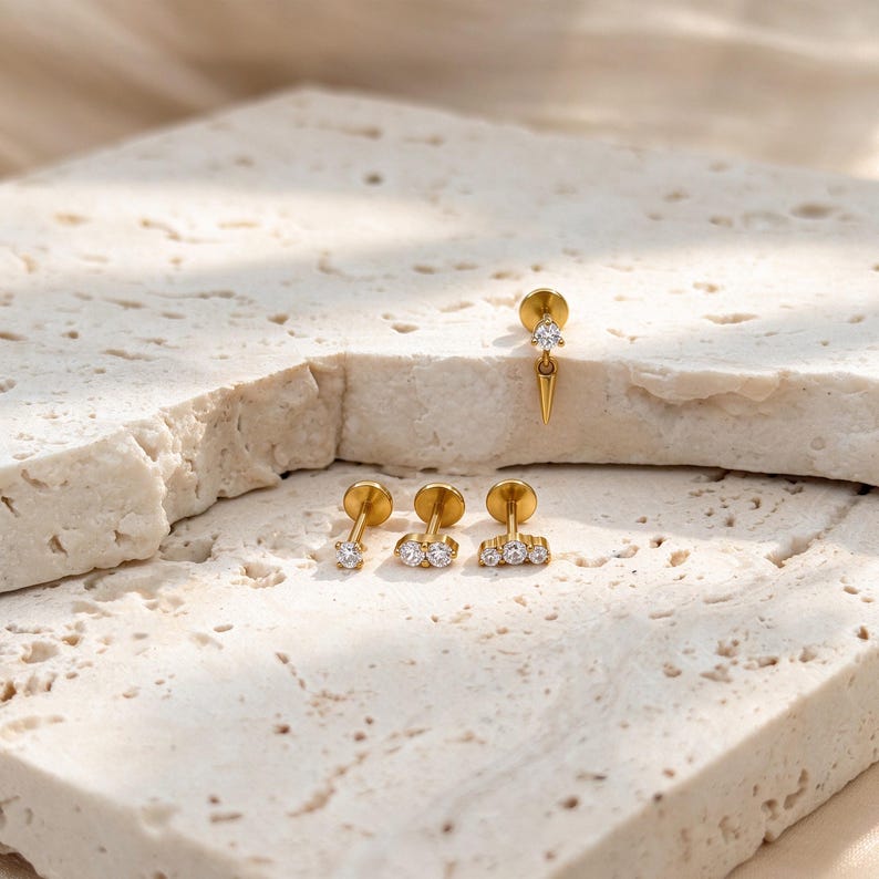 May include: Four gold-tone stud earrings with clear cubic zirconia stones. One earring features a single stone with a dangling spike, while the others are flat studs with one, two, or three stones arranged in a horizontal row on a metal post.