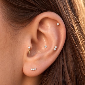Könnte beinhalten: Eine Sammlung goldfarbener Ohrpiercings mit klaren, rund geschliffenen Steinen. Das Set enthält einen spitz zulaufenden Hänger, einen Ohrstecker mit zwei Steinen, einen geschwungenen Stecker mit drei Steinen sowie mehrere Ohrstecker mit einem Stein.