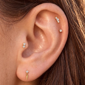 May include: A set of four gold-toned earrings featuring clear, round-cut stones. One earring is a dangling spike, one is a double-stone stud, one is a triple-stone cluster, and one is a single-stone stud, all worn in various ear piercings.