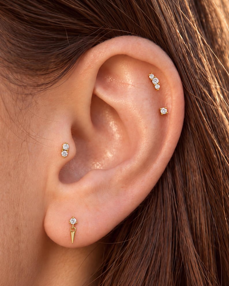 May include: A set of four gold-toned earrings featuring clear, round-cut stones. One earring is a dangling spike, one is a double-stone stud, one is a triple-stone cluster, and one is a single-stone stud, all worn in various ear piercings.