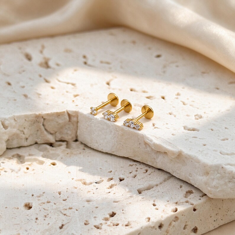 May include: Three gold-tone flat back stud earrings, each featuring a different number of clear, round-cut stones. The first has one stone, the second has two, and the third has three stones arranged in a horizontal row on a metal post.