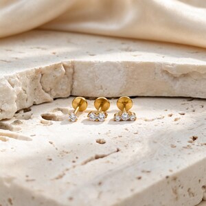 May include: Three gold-tone flat back stud earrings with clear round-cut stones. The first earring features a single stone, the second has two stones arranged horizontally, and the third displays three stones in a curved row, all with metallic posts.