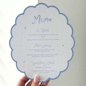 May include: A white menu card with a light blue scalloped edge, featuring handwritten text. The menu lists options for starters, main courses, and desserts. The card includes the words "Menu", "To Start", "Main Course", and "Something Sweet".