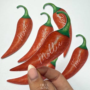 May include: Watercolor illustration of red chili peppers with green stems. Each pepper has a name written on it: Mark, Samantha, Mollie, Rachel, and Toni. The peppers are held by a hand, with the Hills & Ink logo in the bottom left corner.
