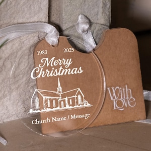 Pode incluir: Enfeite de Natal em acrílico transparente com uma fita branca. O enfeite tem o texto "Merry Christmas" e uma ilustração de uma igreja. Uma etiqueta castanha com "With love" está anexada. Os anos 1983 e 2025 também são visíveis.