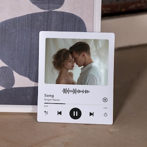 May include: A rectangular white plaque featuring a printed music player interface. The center displays a photograph of two people facing each other. Below the image is a sound wave graphic, the text Song, Singer Name, and playback controls.