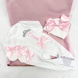 May include: A white baby outfit set with pink accents, including a onesie with the name "Ceiva" and a crown graphic, a matching hat, and booties. The set is adorned with pink bows and presented in a pink gift box.