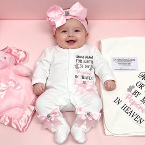 May include: A baby in a white onesie with pink bows and the text "Hand Picked For Earth By My Pepere In Heaven." A matching pink bow headband and a pink plush bunny are visible. A white blanket with the same text is included.