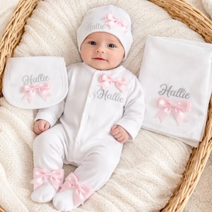 Puede incluir: Un conjunto de ropa de bebé blanco con gorro, babero, body y manta. Cada artículo está adornado con un lazo rosa y el nombre "Hallie" en plata. El body tiene lazos rosas en los pies.