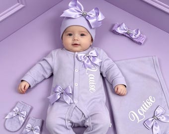 May include: A lavender baby outfit set including a long-sleeved footed sleepsuit with the name Louise embroidered in white, a matching beanie with a large satin bow, a headband, a pair of mittens, and a blanket, all featuring decorative satin bows.