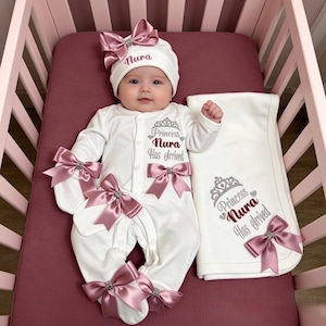 Puede incluir: Un conjunto de bebé blanco con pijama y gorro decorado con lazos de satén rosa y brillantes. El conjunto y una manta blanca a juego incluyen el bordado Princess Nura Has Arrived y un diseño de corona plateada en el tejido.