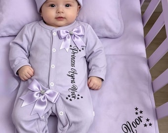 May include: A baby in a lavender outfit, comprising a hat and footed onesie, embellished with satin bows and the name "Ayra Noor." The outfit is on a matching blanket in a cot. The text "Princess Ayra Noor" is printed on the onesie and blanket.
