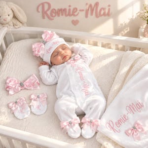 Pode incluir: Um conjunto de bebé branco com macacão, gorro, luvas e manta, decorados com laços de cetim rosa e cristais. O nome Romie-Mai está bordado em rosa. O macacão possui mangas compridas e pés com laços a condizer em cada extremidade.