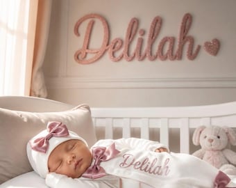May include: A white infant sleeper and matching hat set featuring dusty rose satin bows and the name Delilah in glittery rose gold script. The sleeper has a side-snap closure, and the wall displays the name Delilah in large rose gold letters.