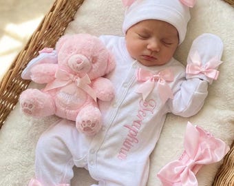 May include: A white long-sleeved footed sleepsuit with pink embroidery reading Seraphina, featuring pink satin bows on the chest, feet, and mittens. Includes a matching white beanie with a large pink bow, a pink headband, and a pink plush teddy bear.