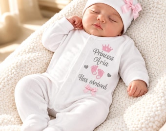 May include: A white long-sleeved baby onesie and matching hat with a pink bow. The onesie features a pink crown, grey text reading Princess Aria, two pink baby footprints, and the text Has arrived, with a small pink bow at the bottom.