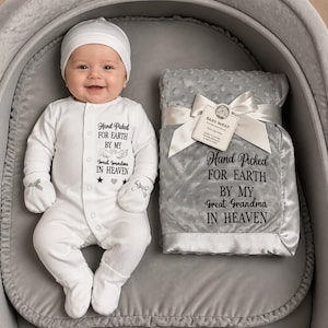May include: A white baby onesie with matching hat and mittens, featuring the text Hand Picked FOR EARTH BY MY Great Grandma IN HEAVEN. Beside it is a grey textured baby blanket with a white satin ribbon and a tag with the same text printed.